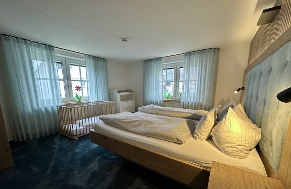 Schlafzimmer mit Doppelbett, Kinderbett links und zwei Fenstern mit hellblauen Vorhängen