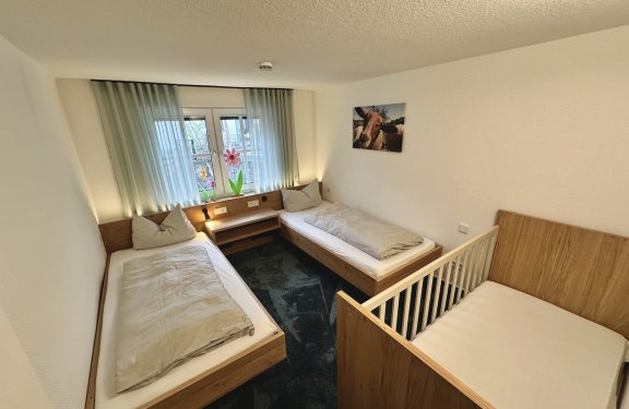 Kleines Schlafzimmer mit zwei Einzelbetten links und rechts, Babybett vorne rechts und Fenster mit Vorhängen