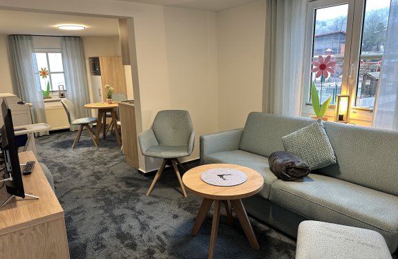 Wohnzimmer mit grauem Sofa, Sessel, kleinem Holztisch und Fenster mit Blumendekoration