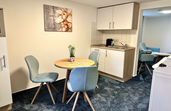 Kleine Küche mit rundem Holztisch, drei blauen Stühlen und einem Bild mit Orchideen an der Wand