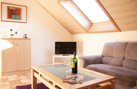 Helles Wohnzimmer mit Dachfenster, Sofa rechts, Glastisch mit Weinflasche und Gläsern, Fernseher und Schrank links