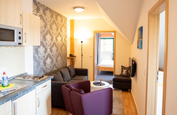 Helle kleine Wohnung mit Küchenzeile links, lila Sessel und Sofa, Blick ins Schlafzimmer