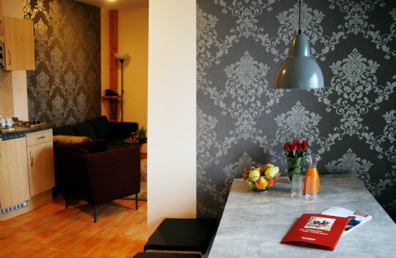 Moderner Wohnbereich mit grauer Tapete, Esstisch mit Obstschale, Blumen und Getränk, angrenzende Küche und Sitzbereich