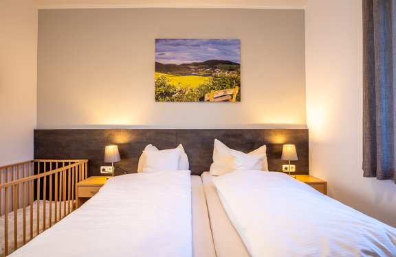 Zwei Einzelbetten mit weißen Bettdecken, Nachttische mit Lampen und ein Babybett links im Zimmer