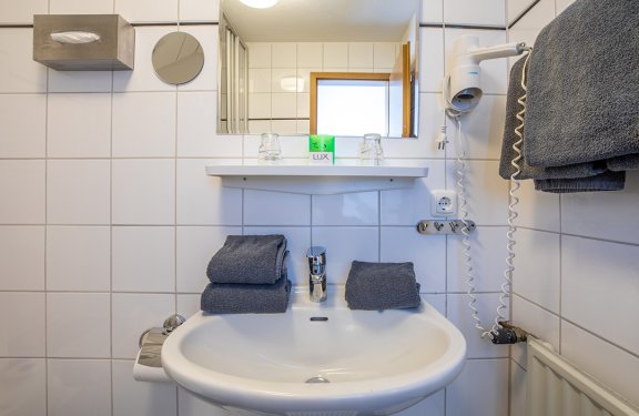 Weißes Waschbecken mit grauen Handtüchern, Spiegel, Föhn und Seife in einem gefliesten Badezimmer