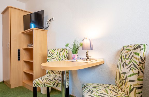 Kleiner Tisch mit zwei gemusterten Stühlen, Lampe und Pflanze an weißer Wand neben Holzschrank mit Fernseher