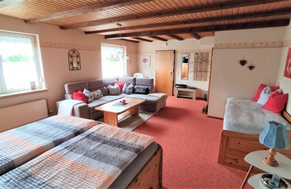 Helles Wohnzimmer mit Holzbalkendecke, grauem Ecksofa, zwei Betten und rotem Teppichboden