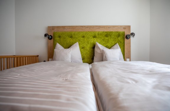 Zwei Einzelbetten mit weißen Bettdecken und Kissen vor einem grünen gepolsterten Kopfteil mit Holzrahmen