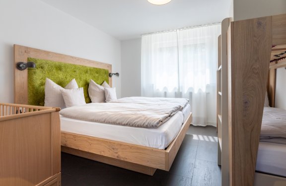 Helles Schlafzimmer mit Doppelbett, grünem Kopfteil, Kinderbett links und Etagenbett rechts