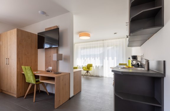 Moderner Wohnraum mit Holzschrank, Schreibtisch, grünem Stuhl, Küchenzeile und Fenster mit Vorhängen