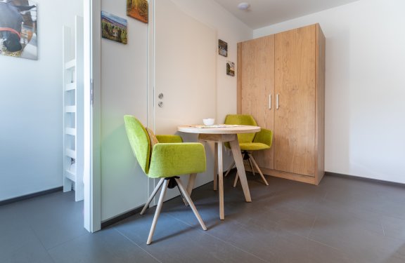 Kleiner Raum mit rundem Holztisch, zwei grünen Stühlen und großem Holzschrank an der Wand