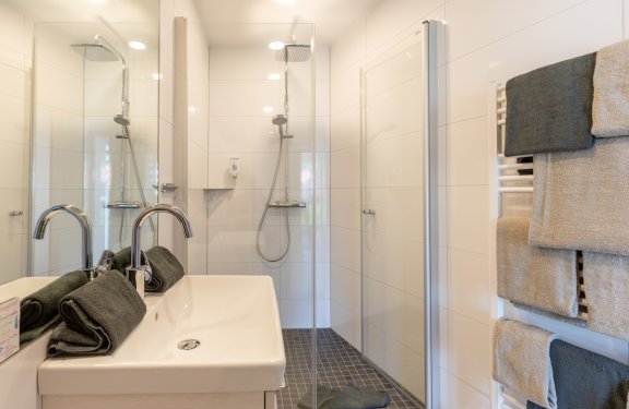 Moderner Badezimmerspiegel links, Waschbecken mit Handtüchern, Dusche in der Mitte, Handtücher rechts am Heizkörper