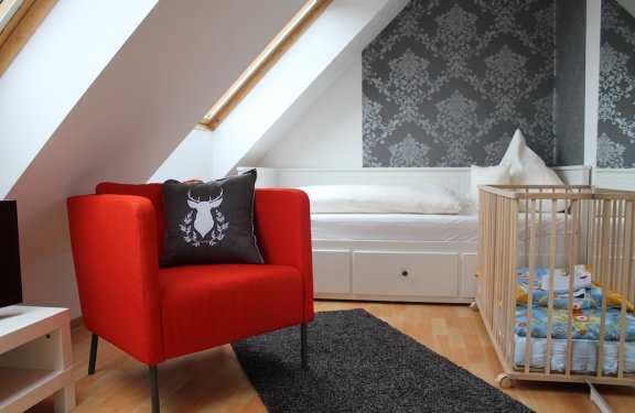 Rotes Sessel mit Hirschkissen vor weißem Bett und Babybett in einem Dachzimmer mit Schrägen