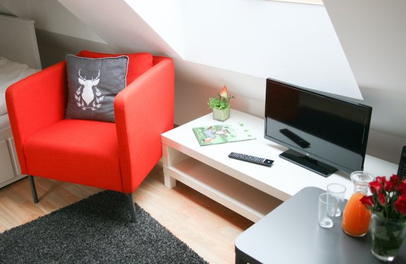 Rotes Sessel mit Hirschkissen neben weißem Tisch mit Fernseher, Fernbedienung und Pflanze unter Dachschräge