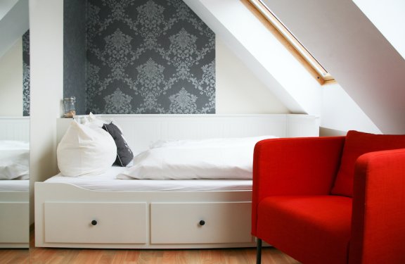 Weißes Bett mit Kissen vor grauer gemusterter Wand und rotem Sessel unter Dachfenster